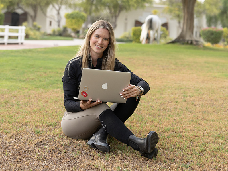 tcc_caro_laptop "Eine lächelnde Frau in Reitkleidung sitzt mit einem Laptop im Gras, während im Hintergrund ein Pferd grast.